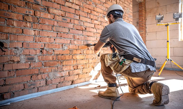 Hombre con casco sentado con un taladro en una pared de ladrillo
